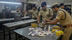 Fake Toothpaste Factory Busted in Delhi, Owner Arrested Police officers inspecting a small illegal manufacturing unit where workers are filling counterfeit toothpaste into branded tubes, with boxes of fake packaging and seized materials visible around the room.