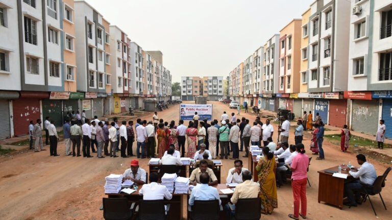 A government notice document displaying details of a public auction for commercial shops under the Telangana 2BHK housing scheme, including a table listing locations, shop sizes, prices per square foot, and auction dates.